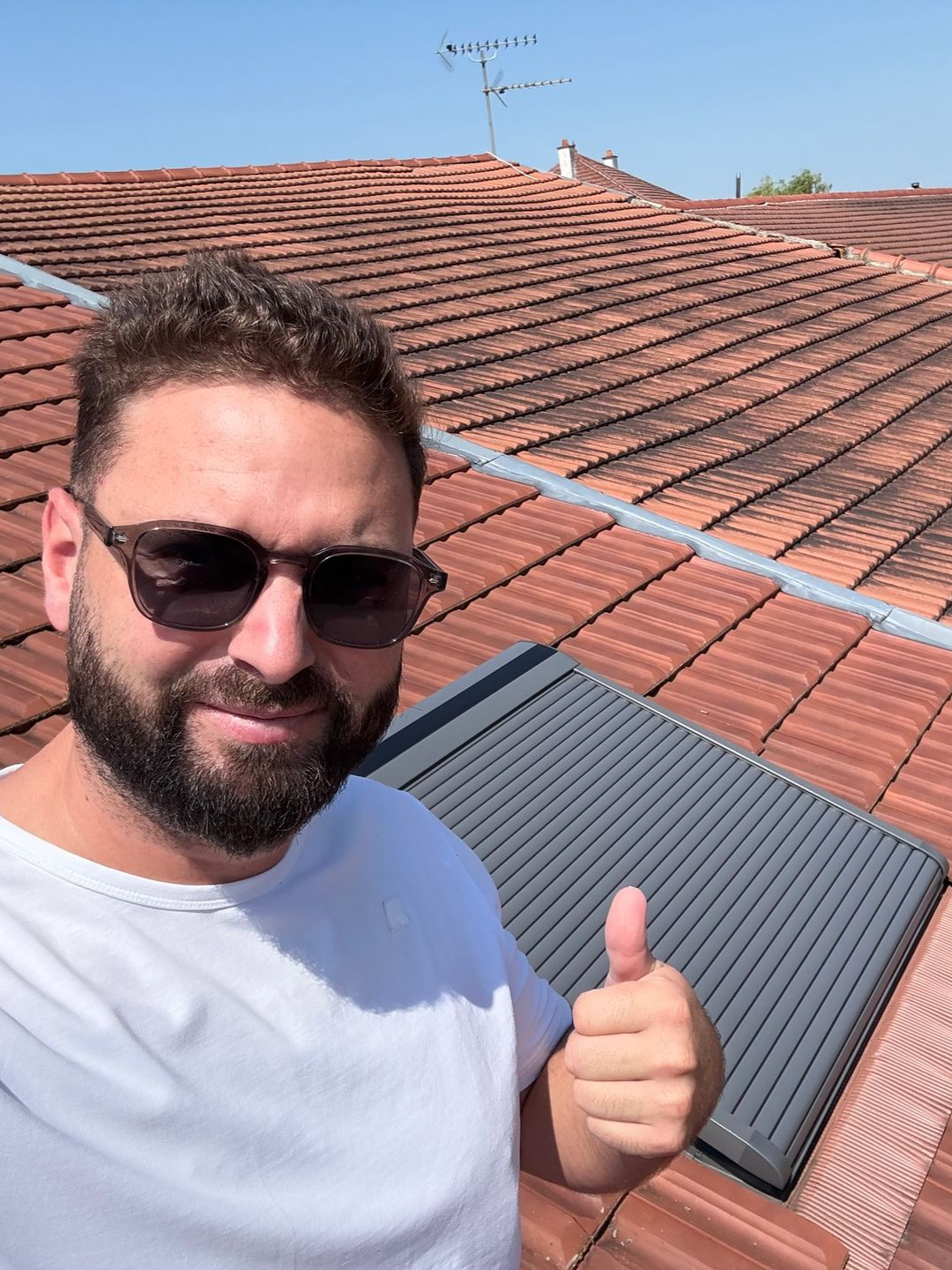 Un homme sur un toit, pouce levé, près d'un panneau solaire. Toit de tuiles rouges, ciel bleu.