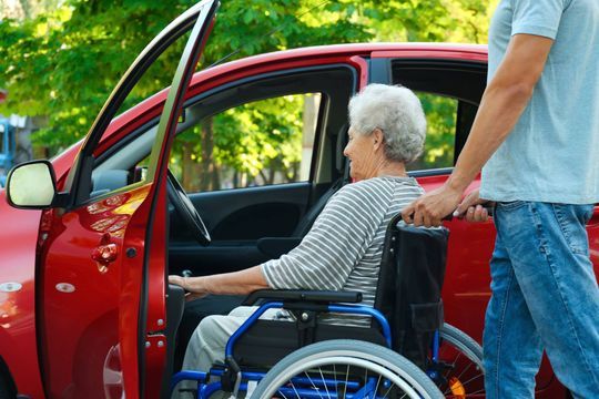 Ältere Frau im Rollstuhl wird zu einem roten Auto hingefahren
