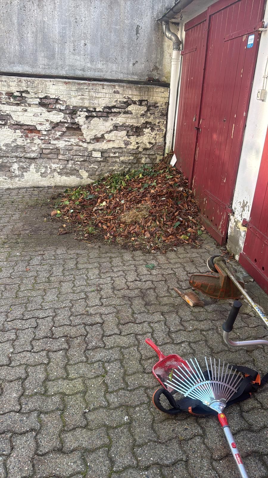 Laubhaufen nahe einer Backsteinmauer und roten Türen; ein Rechen liegt auf einem Kopfsteinpflaster.