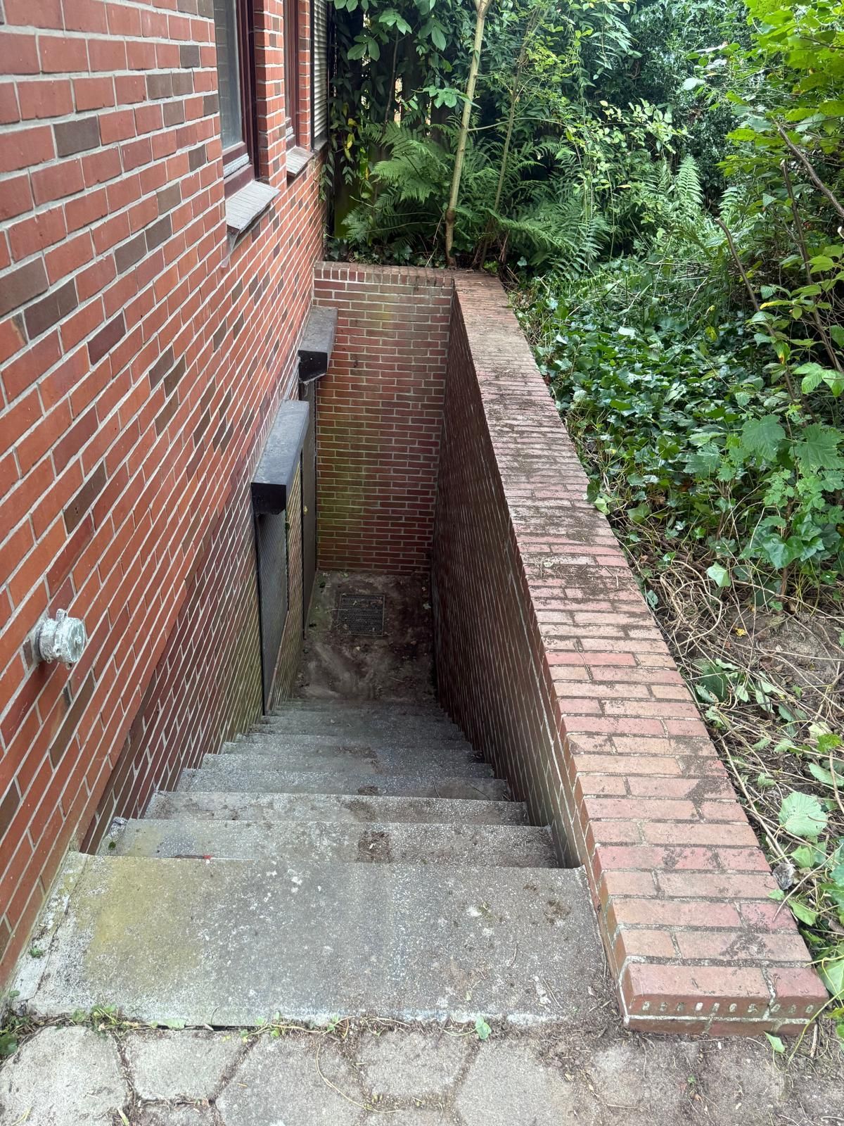 Ein gemauertes Treppenhaus führt hinunter zu einem zurückgesetzten Türeingang; rechts davon befinden sich Ziegelwände und Laubwerk.