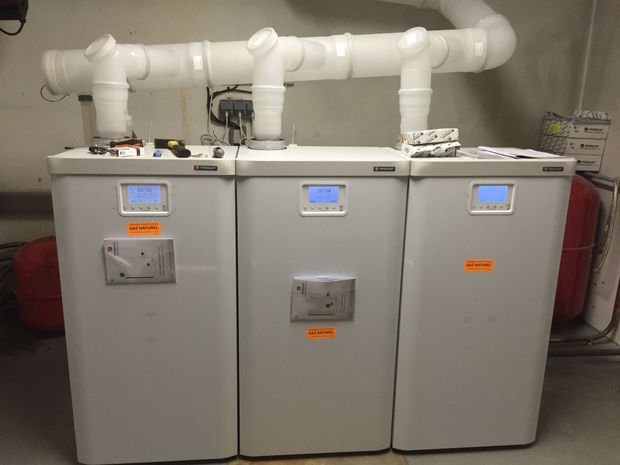 Trois unités de chauffage grises avec tuyaux blancs dans un sous-sol. Affichages numériques sur les unités.