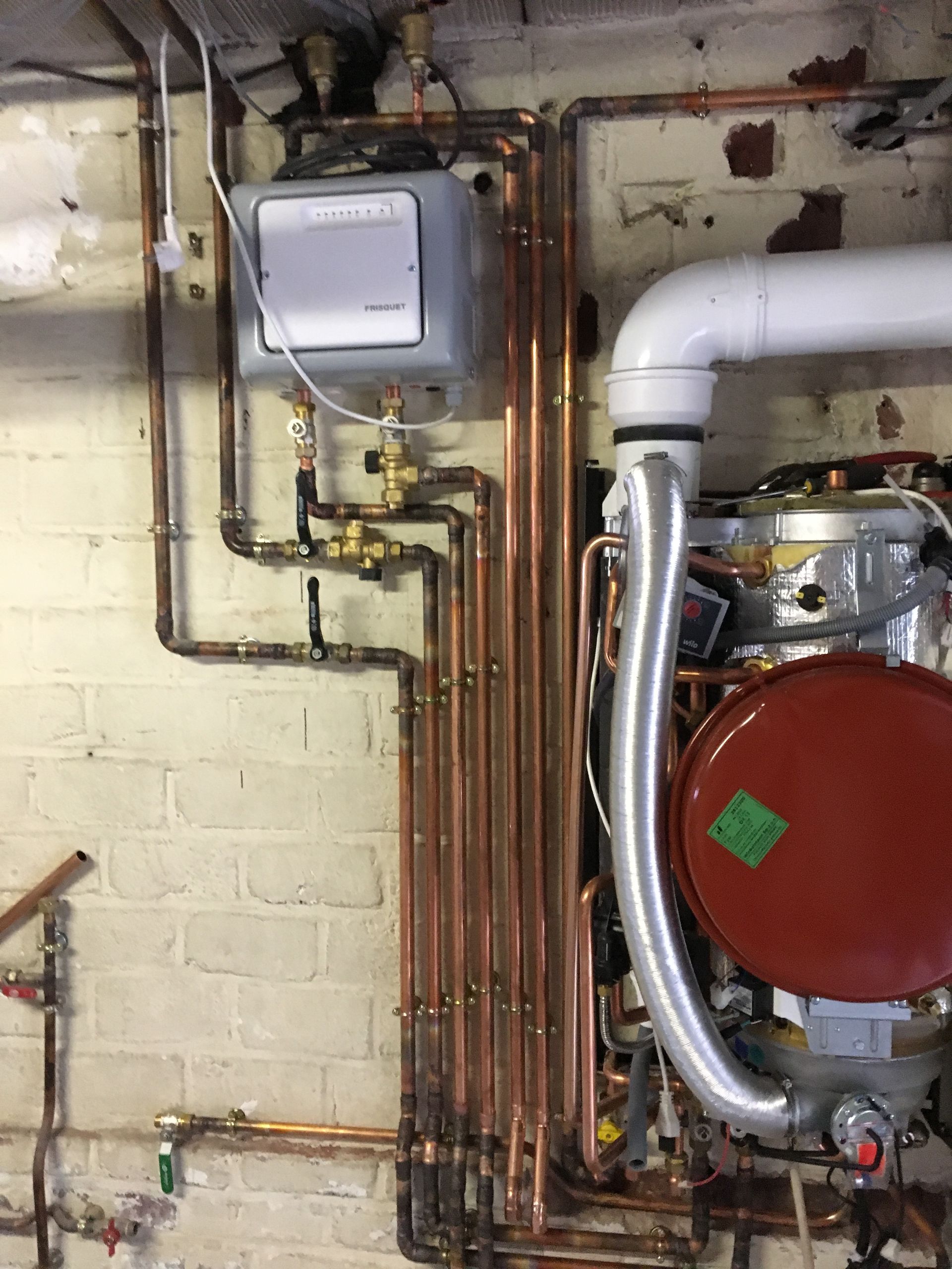 Des tuyaux en cuivre et une chaudière sont reliés à une boîte grise, un réservoir rouge et d'autres équipements contre un mur de briques.