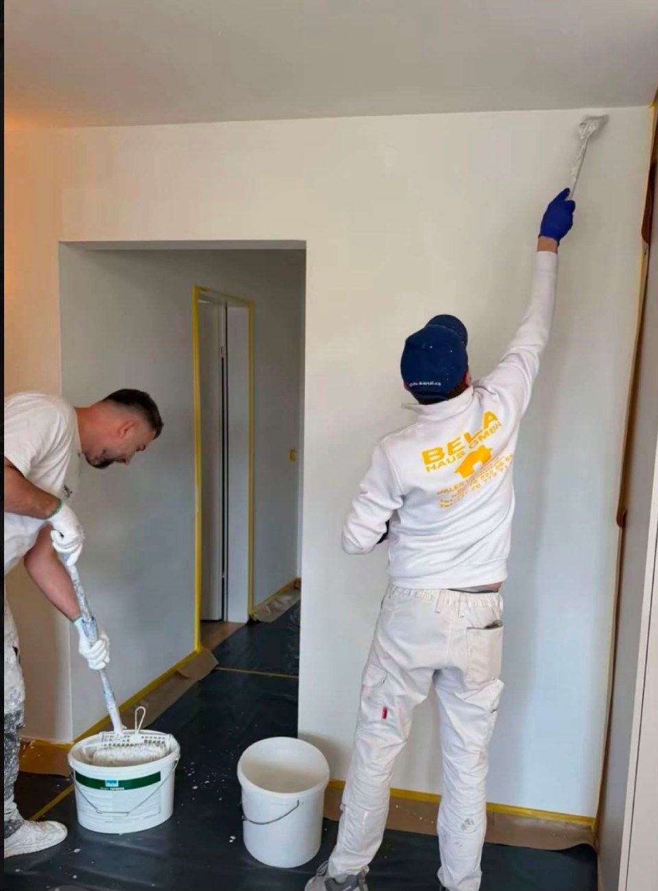 Ein Maler in blauem Overall und Schutzhelm trägt in einem hellen Raum gelbe Farbe auf eine weisse Wand auf.