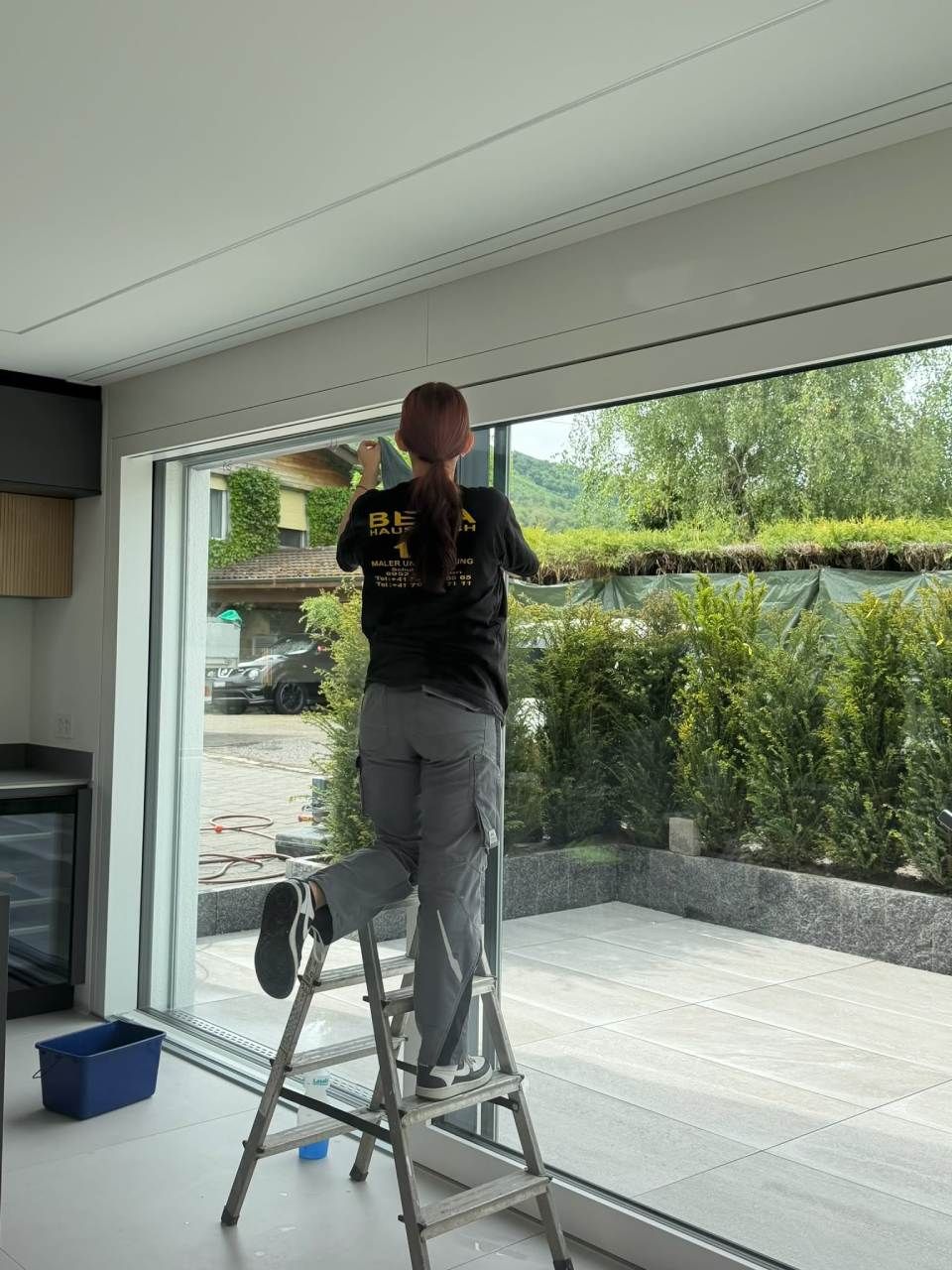 Ein Arbeiter montiert auf einer Trittleiter Fensterverkleidungen in einem Raum mit grossen Glastüren.