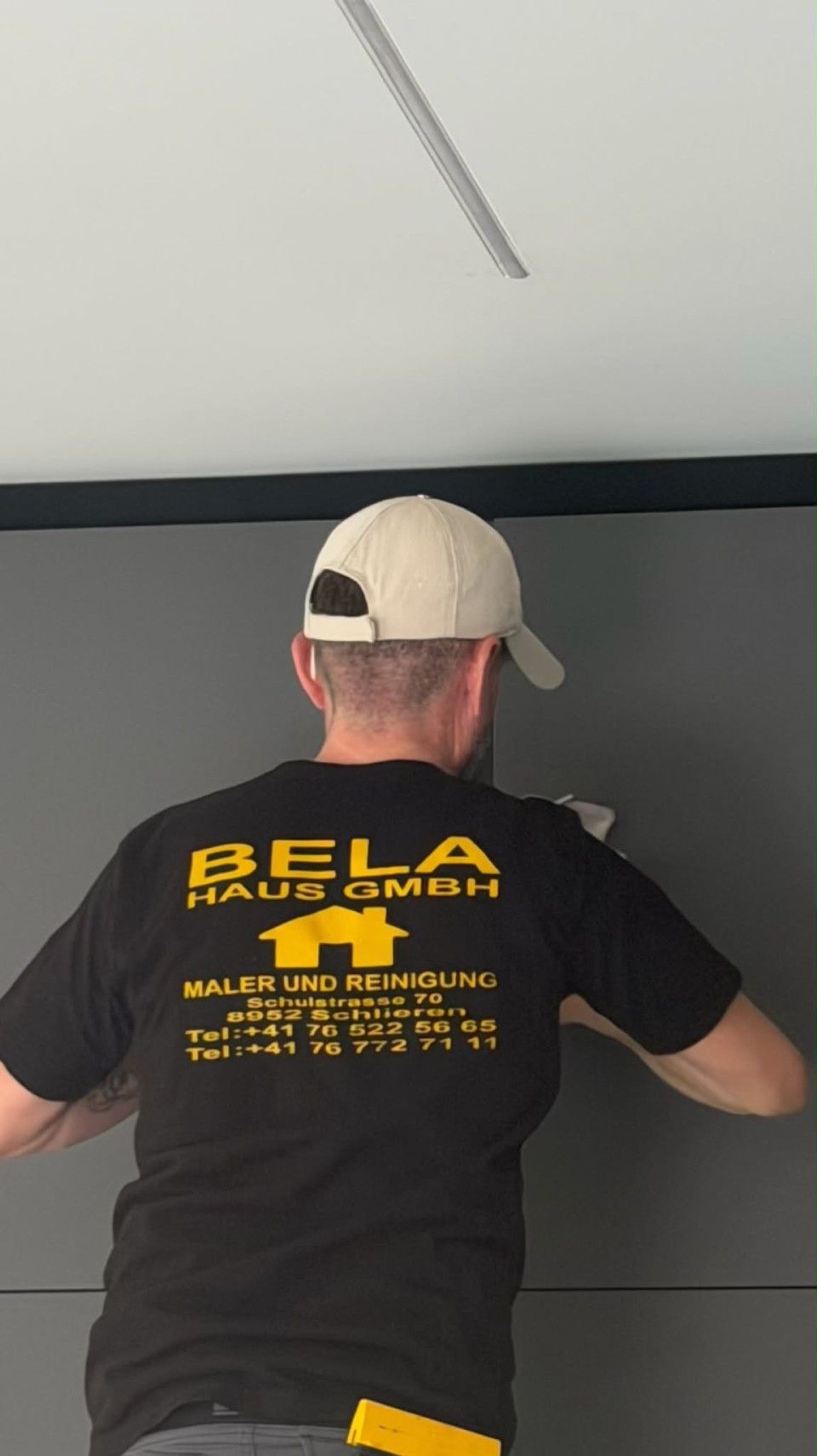 Mann in einem schwarz-gelben BLA-Shirt und beiger Kappe, von hinten gesehen, arbeitet an einer Wand in einem Innenraum.