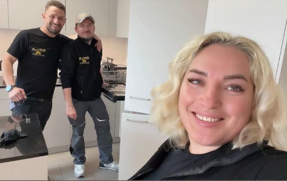 Drei Personen lächeln in einer Küche, zwei stehen an der Küchentheke und eine macht ein Selfie.