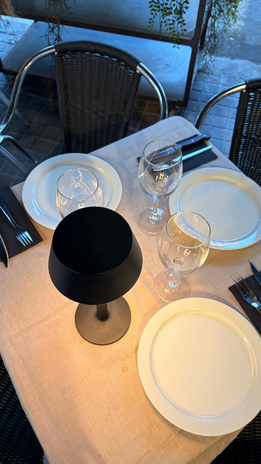 Mesa puesta con platos blancos, copas de vino y una lámpara negra sobre un mantel beige, junto a una ventana