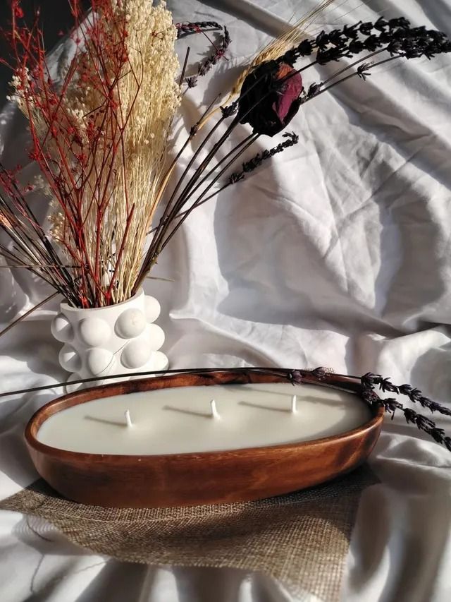 Una vela en un recipiente de madera está sobre una mesa al lado de un jarrón de flores secas.