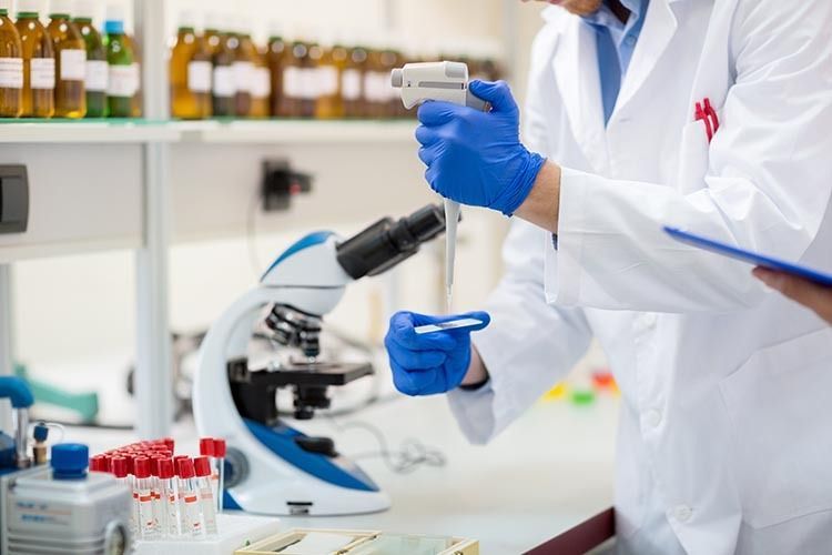 Científico en un laboratorio usando un microscopio y guantes azules, manipulando muestras cerca de tubos de ensayo y frascos.
