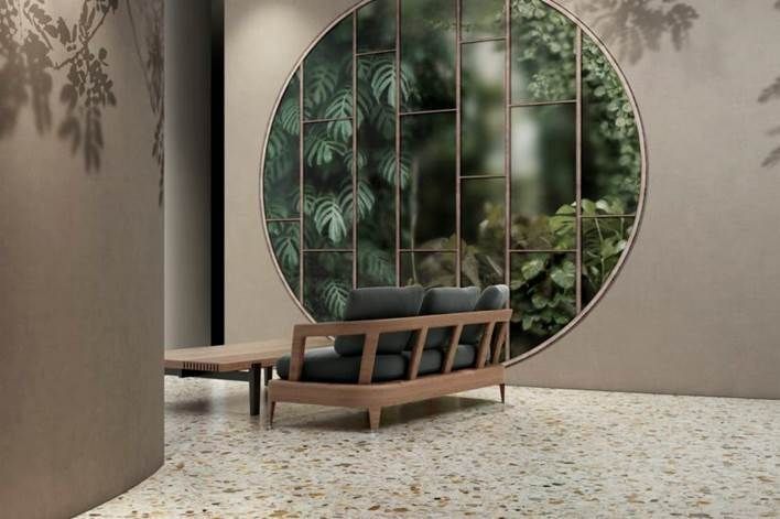 Una moderna sala de estar con una ventana redonda que exhibe exuberantes plantas detrás de un sofá con marco de madera y una mesa de café, piso de terrazo y colores neutros.