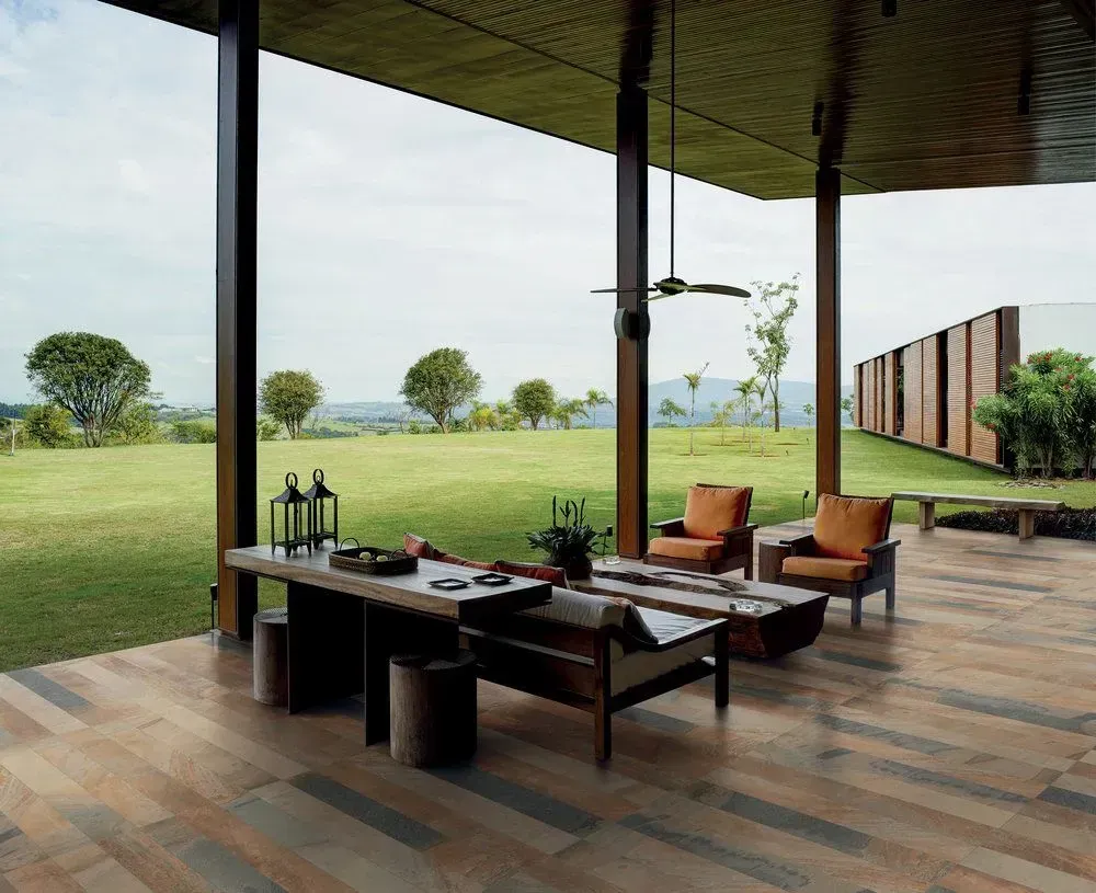 Un patio cubierto con muebles de madera da a un amplio césped verde con árboles y una valla de madera moderna en la distancia.