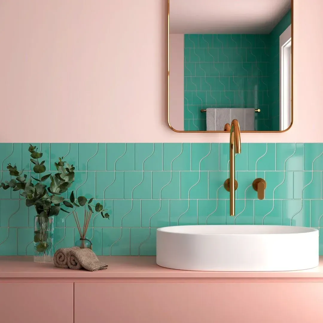 Baño con paredes y tocador de color rosa, salpicadero de azulejos color verde azulado, lavabo redondo blanco, grifo dorado y espejo rectangular.