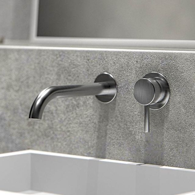 Grifo de baño de pared de acero cepillado con manija de palanca, colocado sobre una pared gris texturizada y un lavabo blanco.
