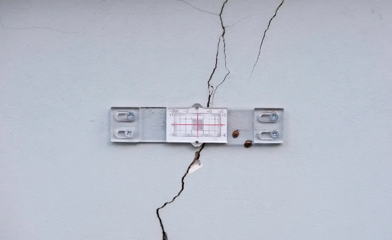 Graue Wand mit einem großen Riss, eine Metallplatte mit Gitterlinien und ein kleiner Rissindikator.