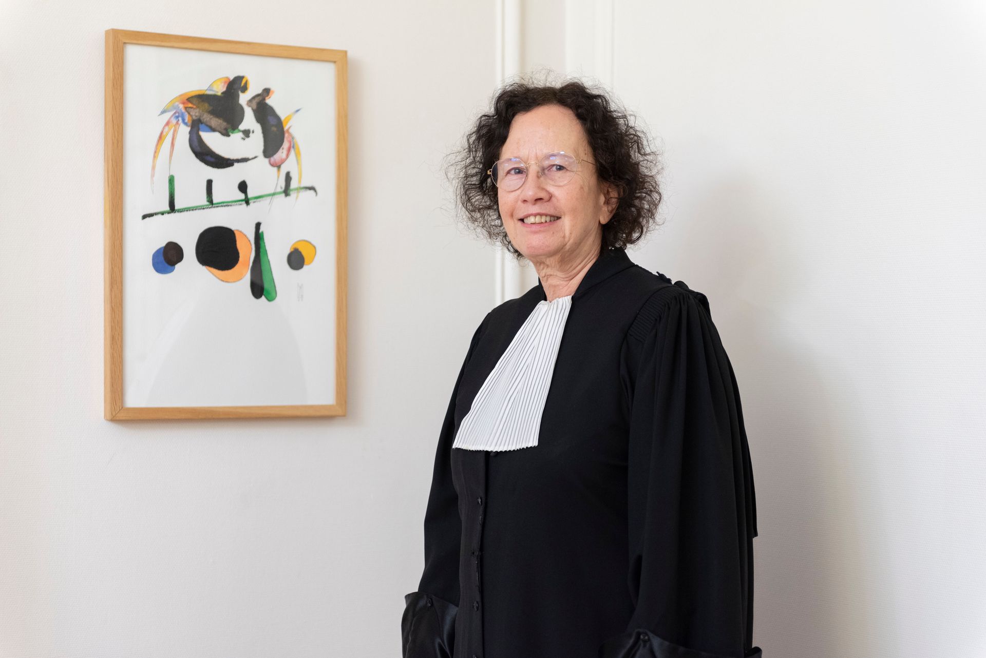 Une femme en veste noire se tient à côté d'un tableau abstrait encadré, accroché à un mur blanc.