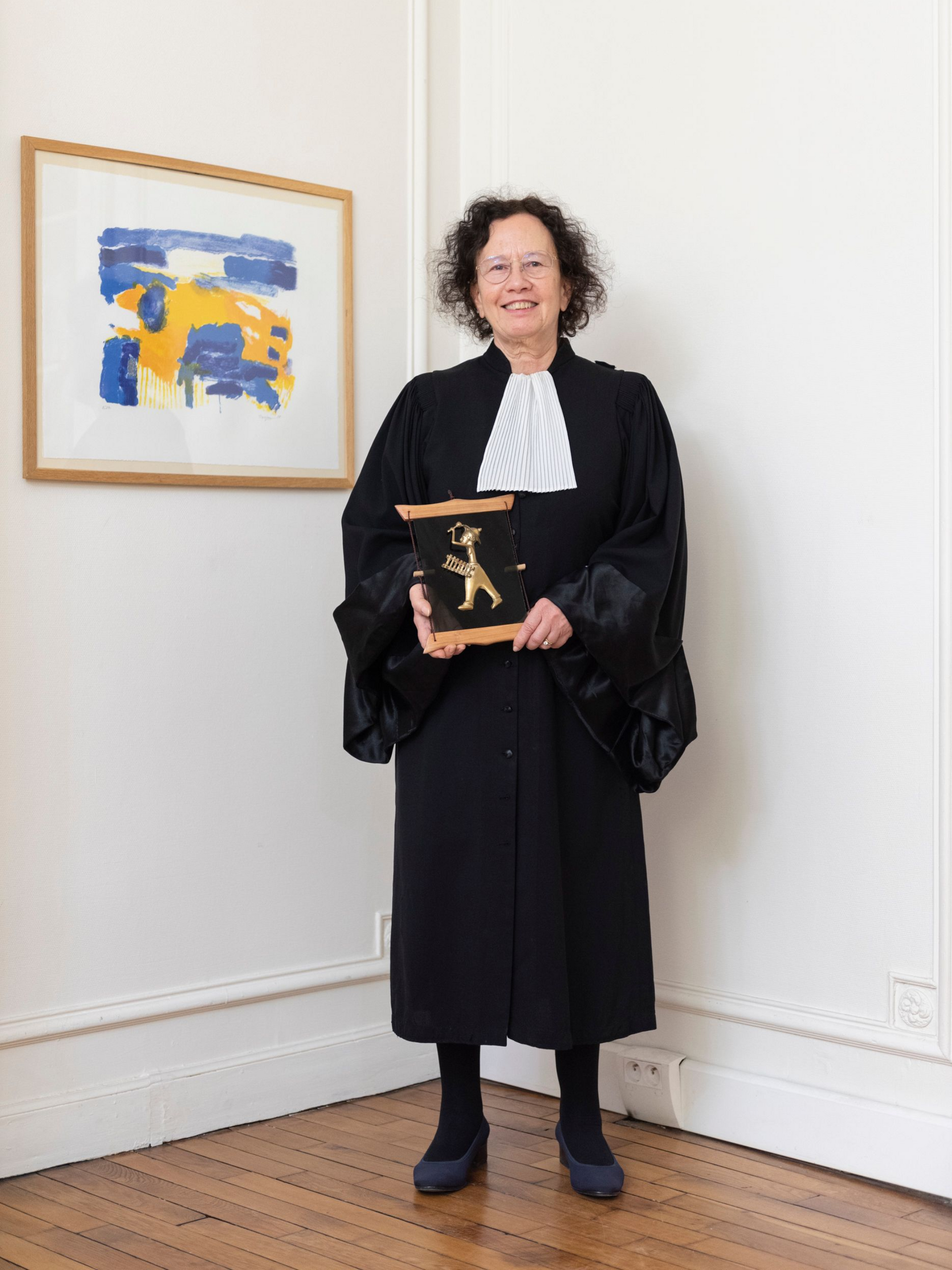 Une personne en robe noire, tenant un prix, se tient dans une pièce lumineuse à côté d'une œuvre d'art encadrée.