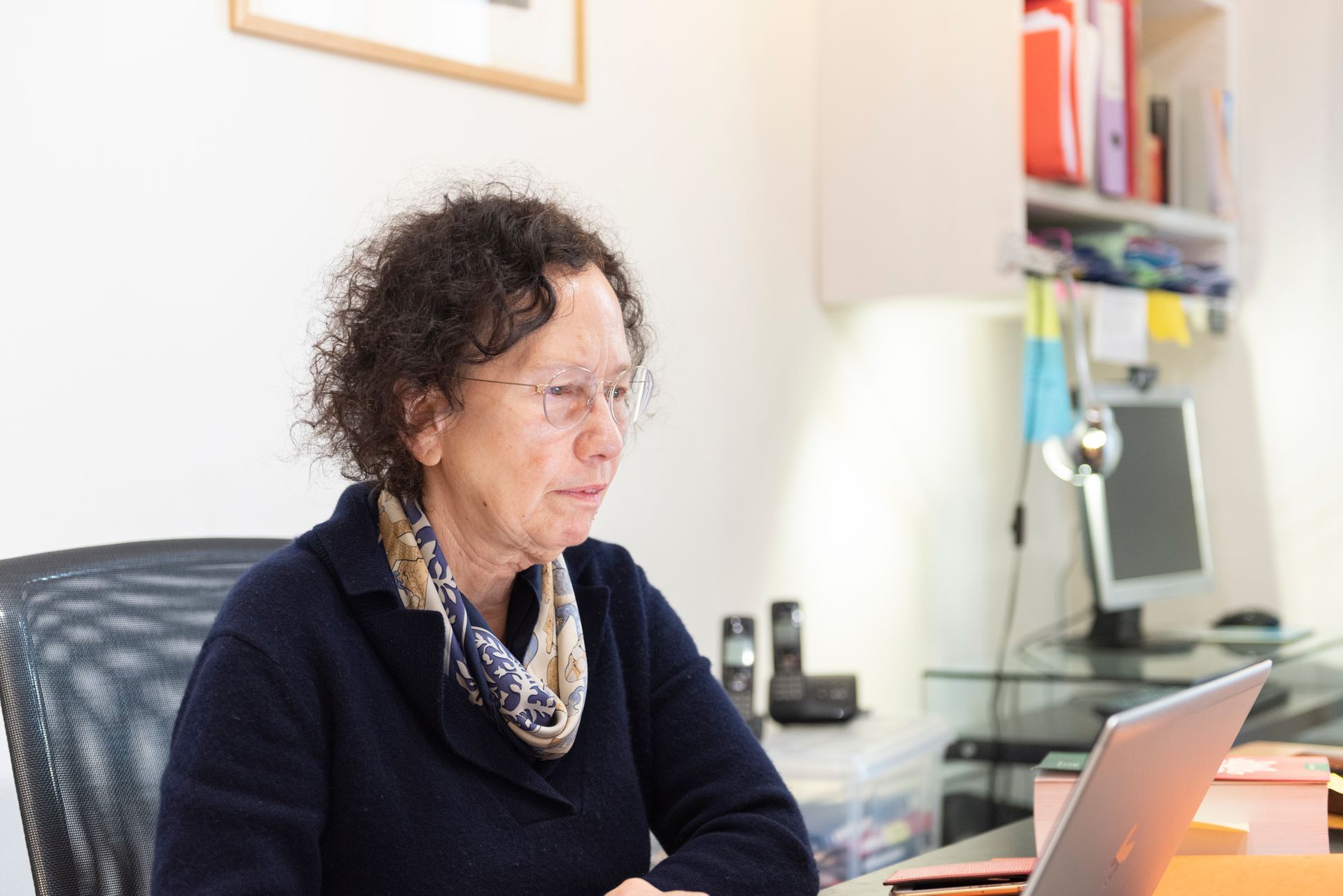 Une personne travaillant à un bureau dans un bureau, regardant un ordinateur portable avec des papiers et des étagères en arrière-plan.
