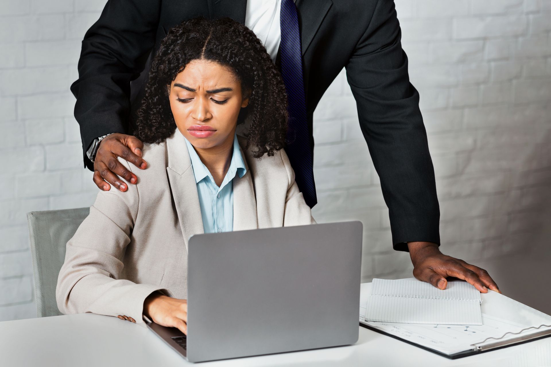 Un employé de bureau stressé, les yeux rivés sur son ordinateur portable, tandis qu'un collègue en costume se tient derrière lui, la main sur l'épaule, dans une salle de réunion.