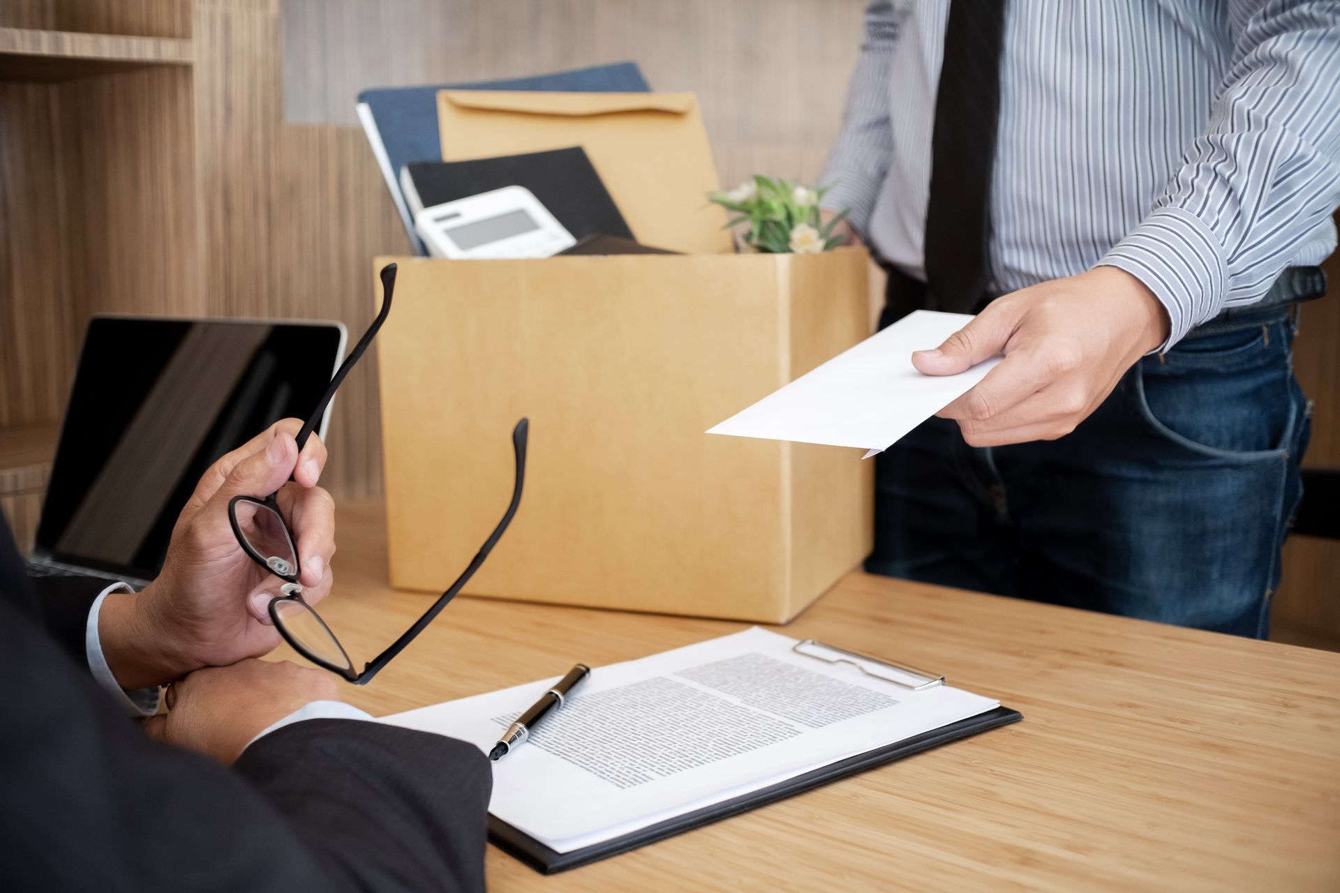 Une personne remet une enveloppe par-dessus un bureau, un ordinateur portable et des lunettes à la main, dans un environnement de bureau.
