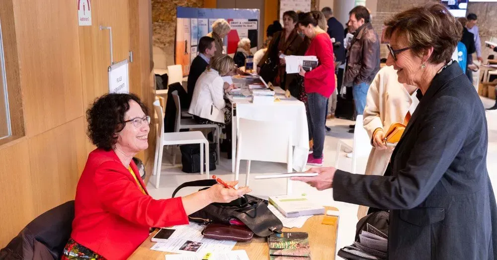 Une femme en veste rouge discute avec une autre femme à une table d'inscription dans une salle événementielle très fréquentée.