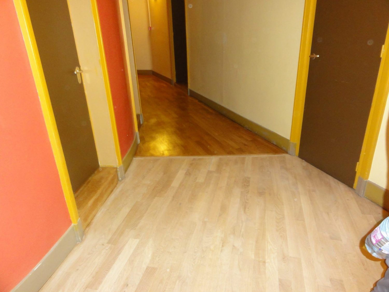 Un couloir avec un parquet clair, des portes marron à encadrement jaune et des touches de couleur orange aux murs, menant à un couloir en bois.