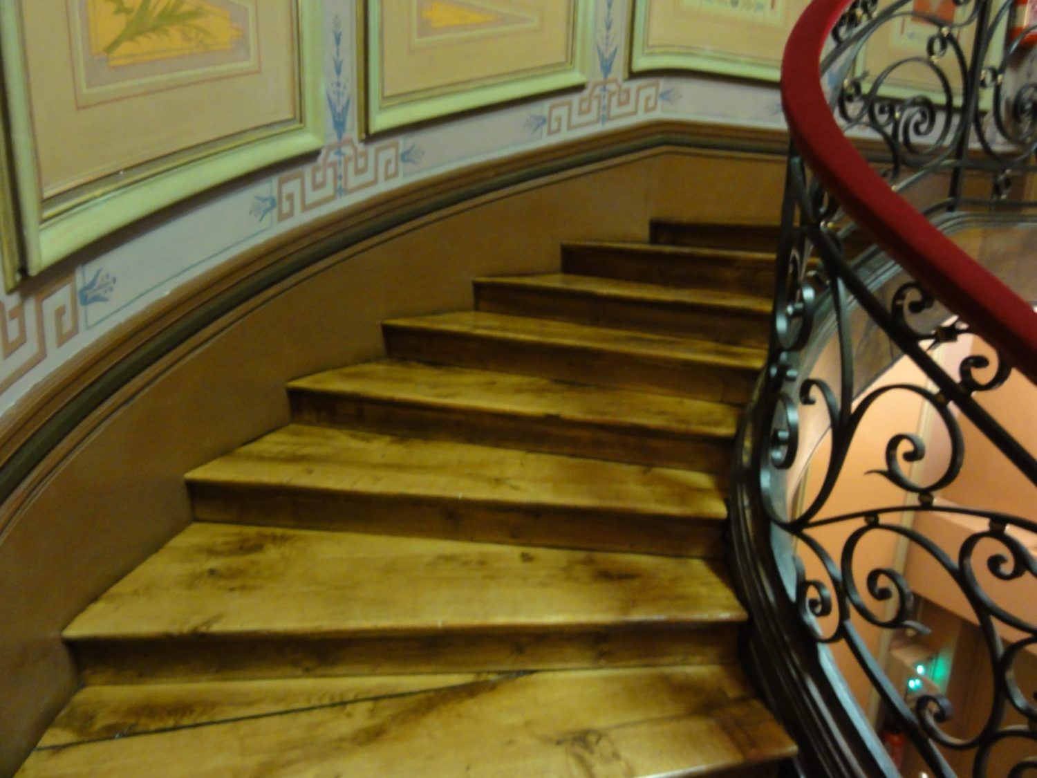 Escalier en bois courbé avec une rampe en fer forgé noir ornée et une main courante rouge, adossé à un mur décoré d'œuvres d'art encadrées.