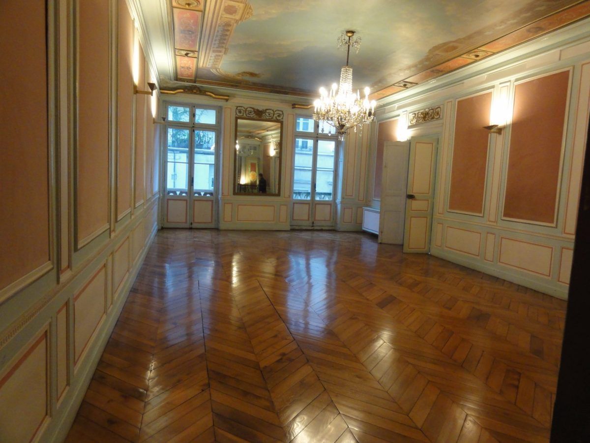 Une pièce vide et richement ornée, avec des murs clairs, un parquet en bois, un lustre en cristal et de grandes fenêtres.