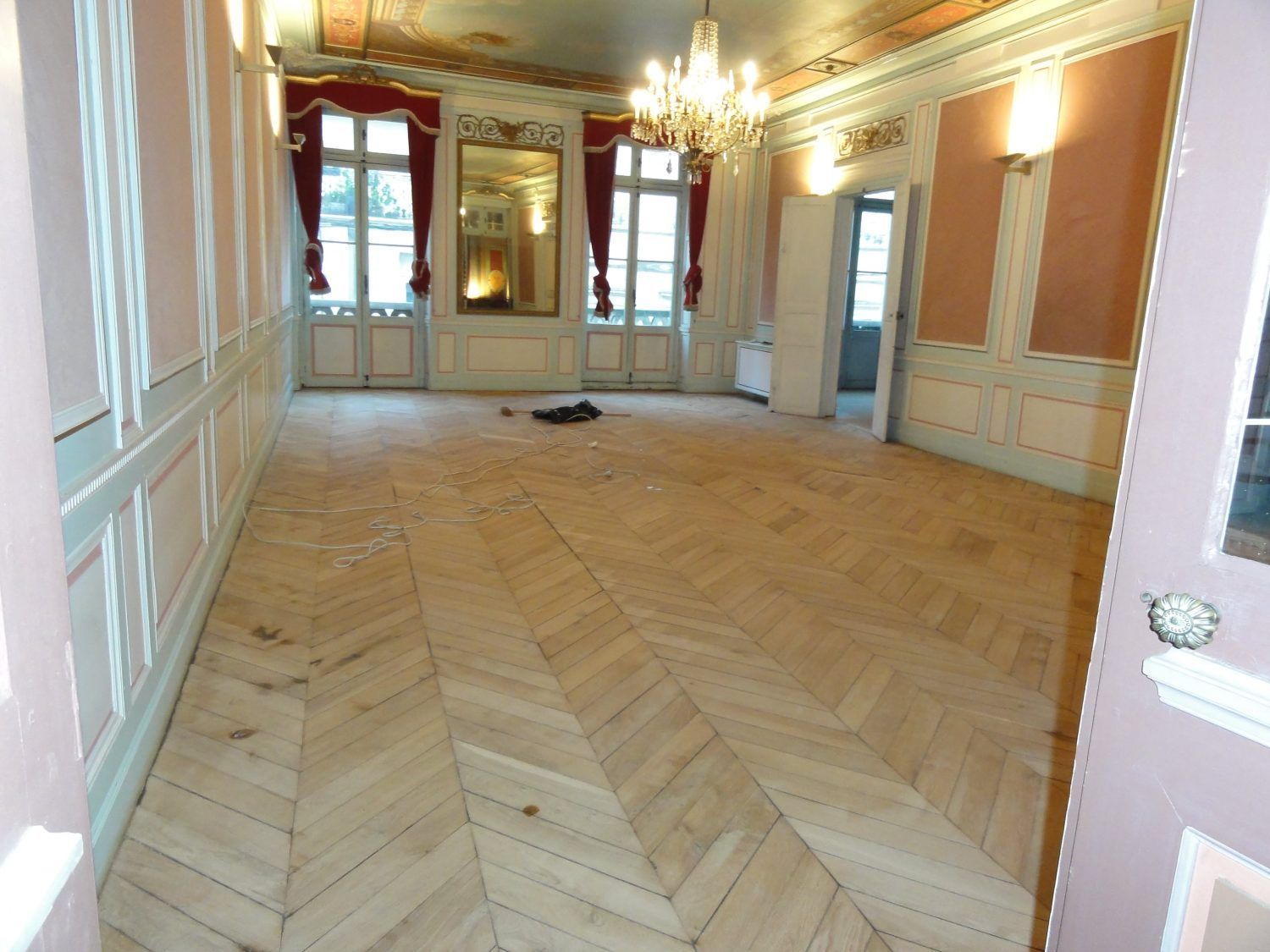 Une pièce vide aux murs rose pâle, aux lambris blancs, au parquet clair à chevrons et au grand lustre.