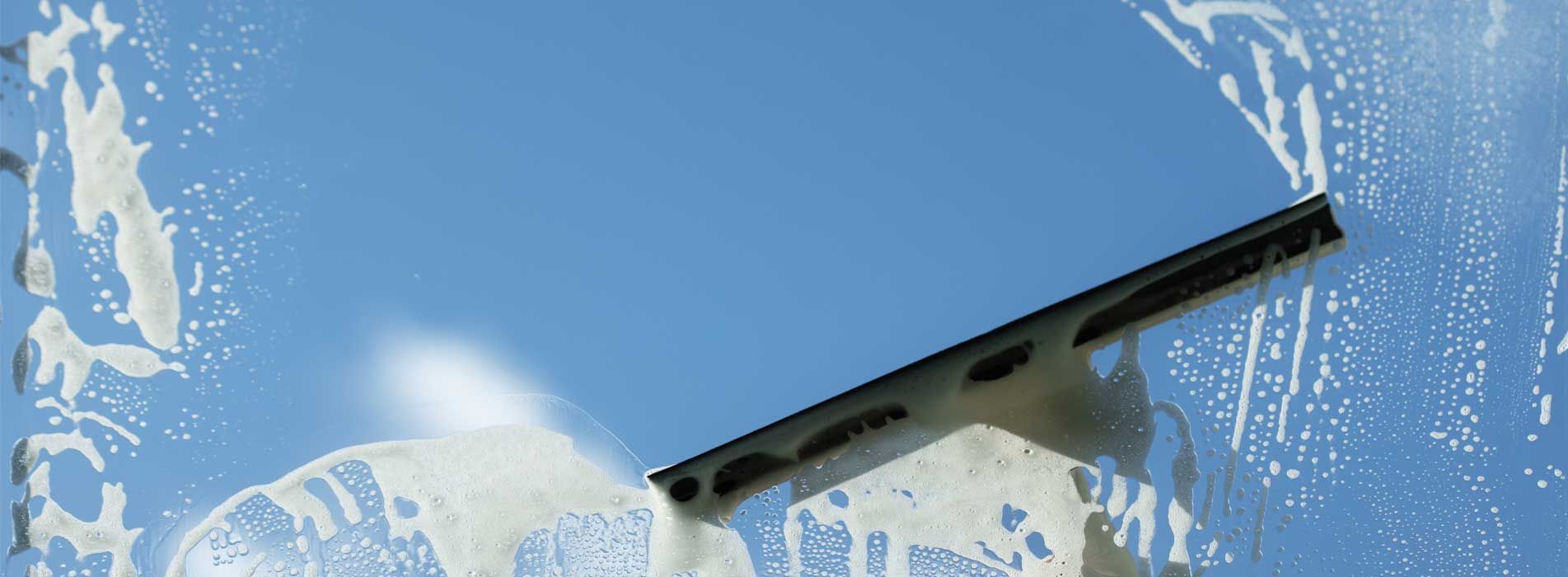 Une raclette essuie la mousse de savon sur une vitre, révélant un ciel bleu limpide derrière elle.