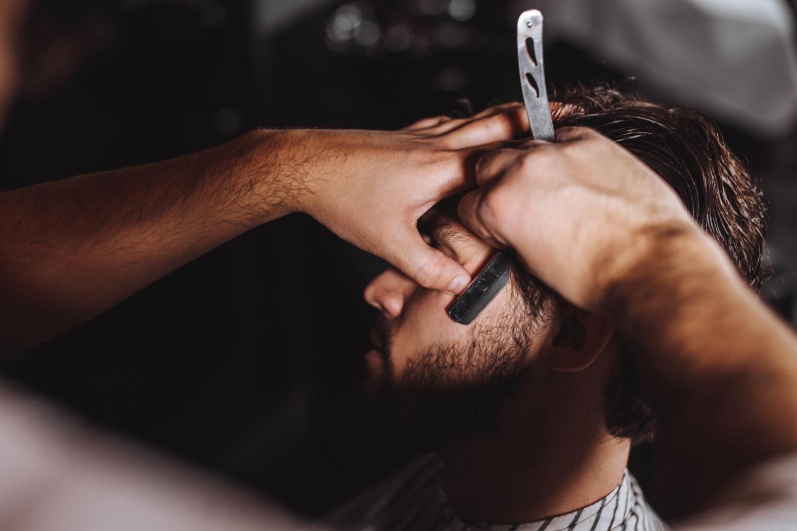 Barbero afeitando la mejilla de un cliente con navaja. Primer plano en una barbería con poca luz.