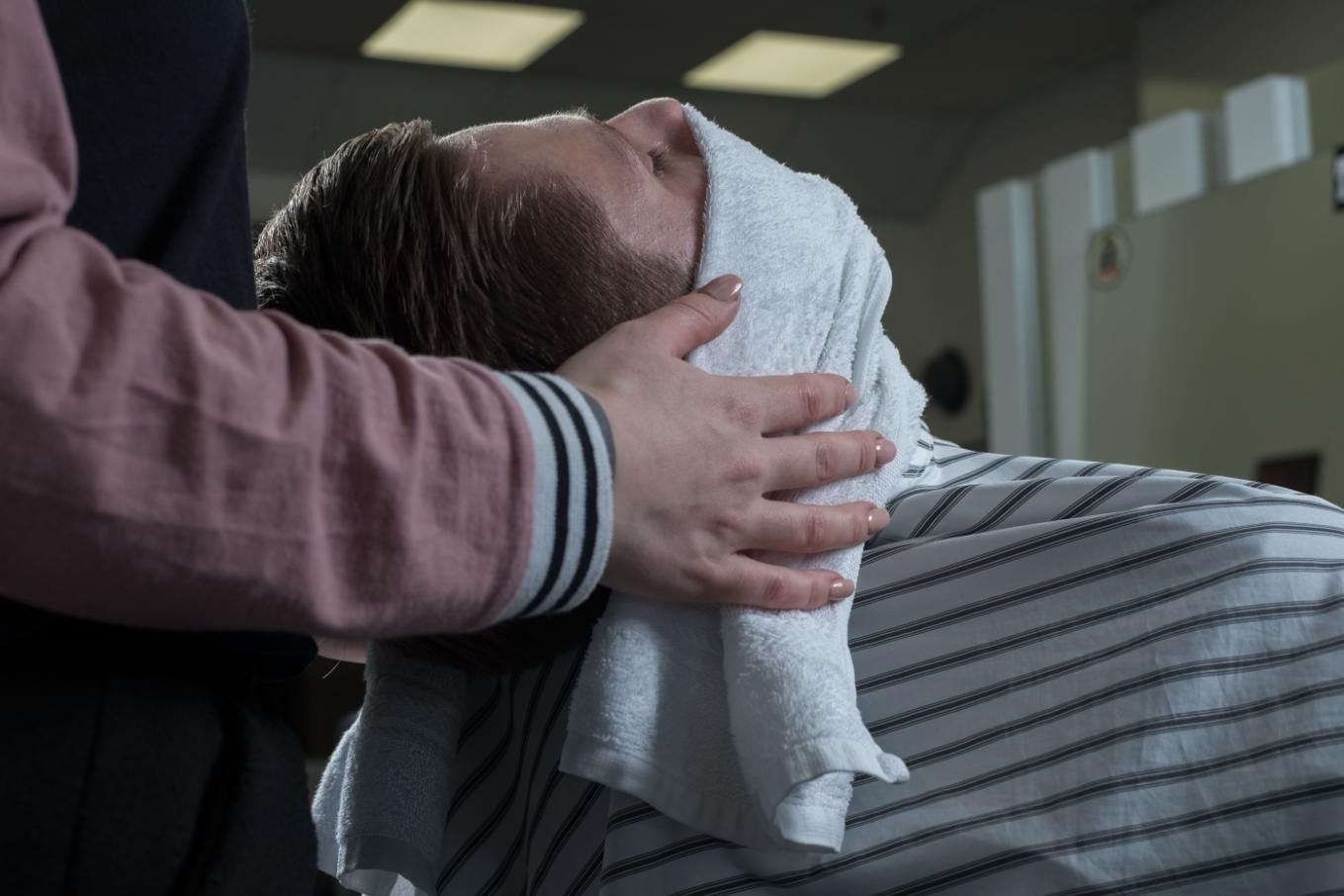 Una persona en una silla de barbero tiene una toalla caliente en su cara, sostenida por otra persona.