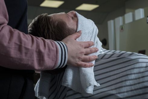 Una persona en una silla de barbero tiene una toalla caliente en su cara, sostenida por otra persona.