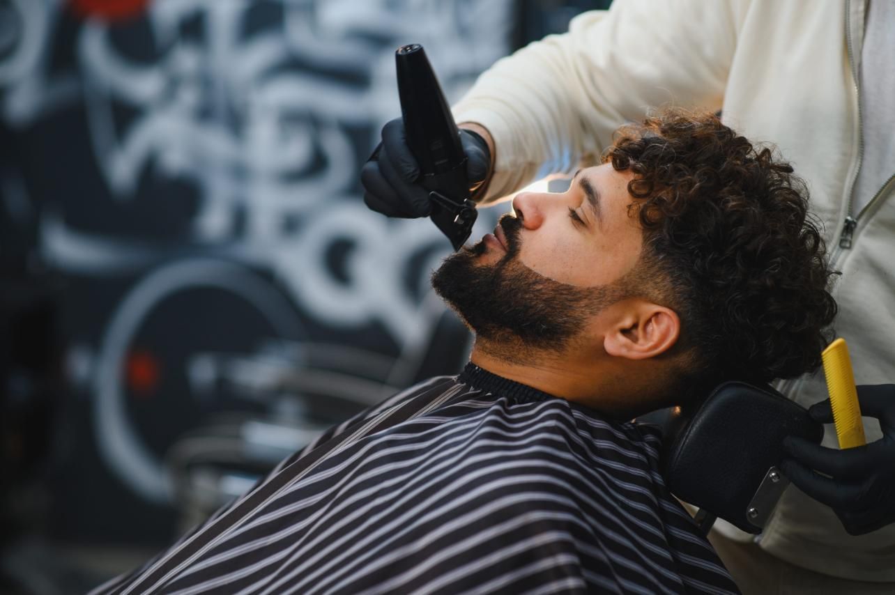 Un barbero recorta la barba de un cliente con una recortadora. El cliente está sentado en una silla, con una capa de barbero.