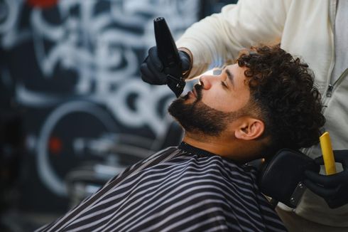 Un barbero recorta la barba de un cliente con una recortadora. El cliente está sentado en una silla, con una capa de barbero.