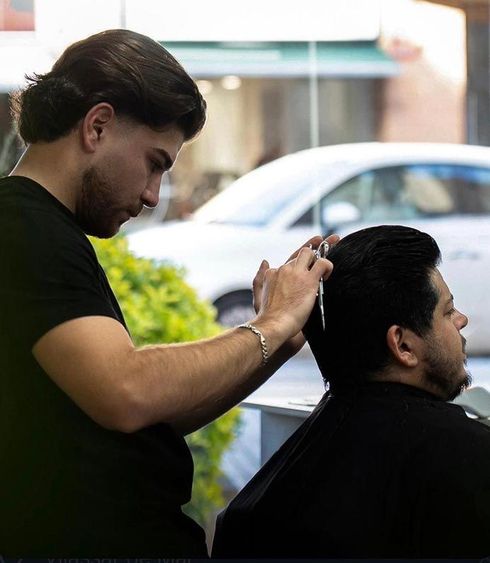 Un barbero le está cortando el pelo a un cliente en una peluquería, con un coche visible fuera de la ventana.
