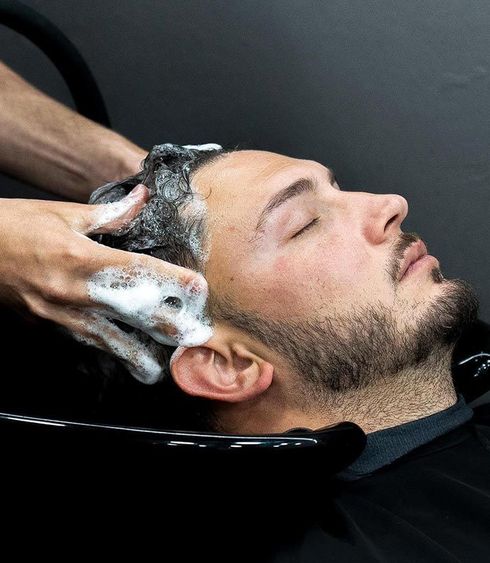 Persona recibiendo un champú en el lavabo de una peluquería, con el pelo enjabonado y los ojos cerrados.
