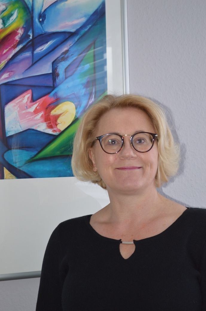 Eine Frau mit Brille lächelt vor einem farbenfrohen abstrakten Gemälde. Sie trägt ein schwarzes Oberteil.