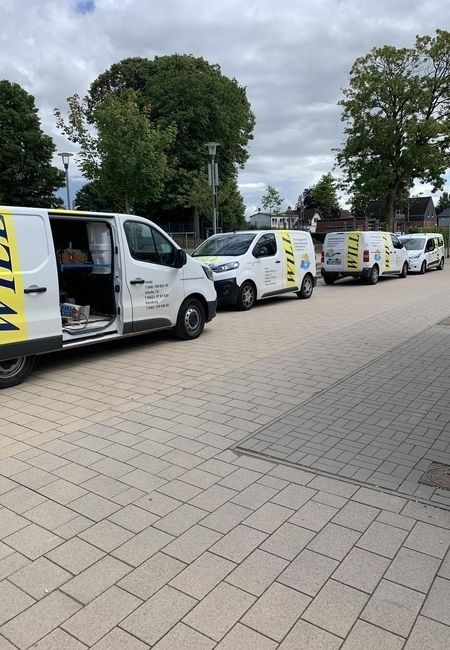 Weiße Lieferwagen standen in einer Reihe auf einer gepflasterten Straße neben Bäumen, bei einem Wagen war die Seitentür geöffnet.