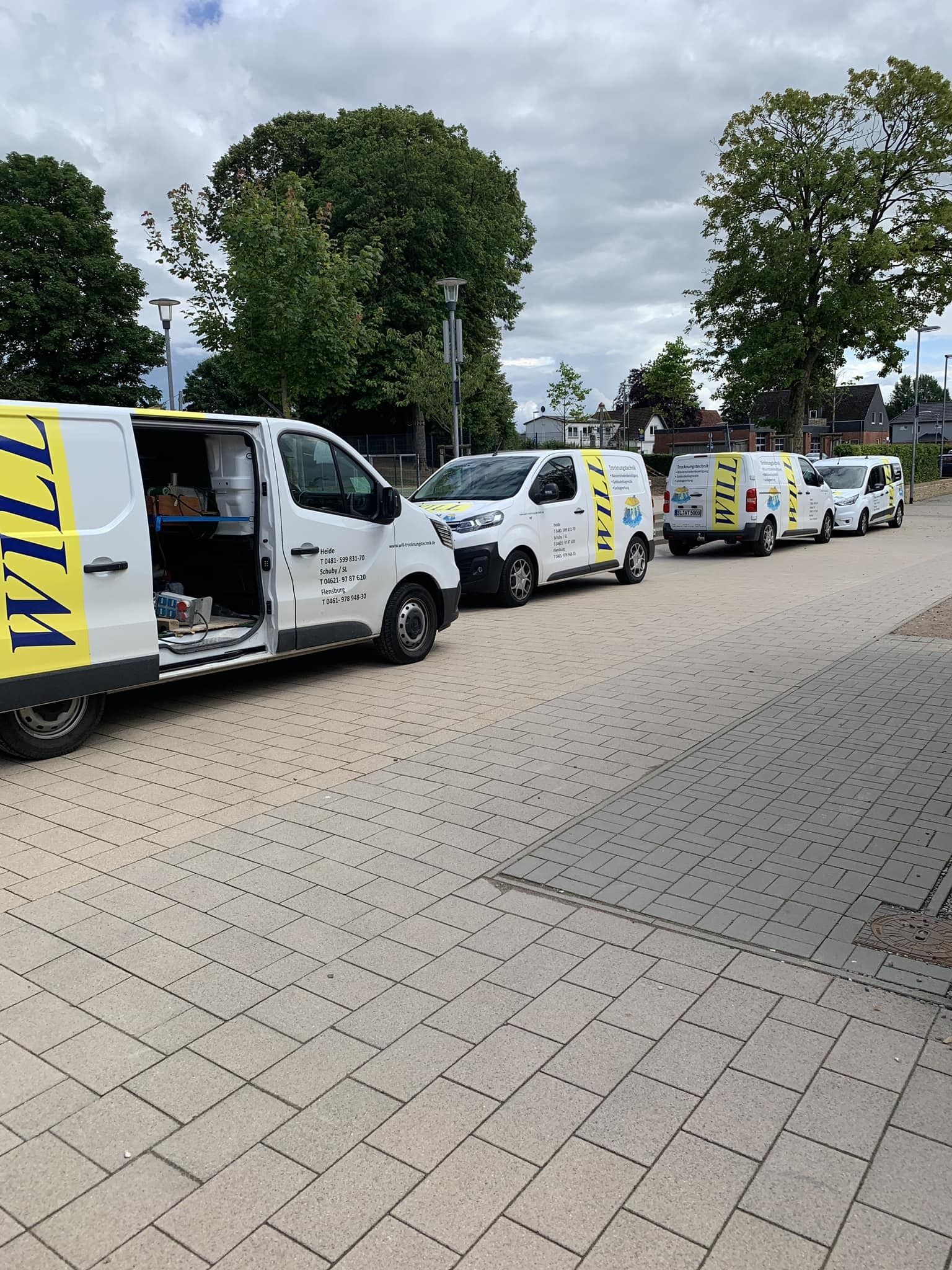Weiße Lieferwagen parken in einer Reihe auf einem gepflasterten Parkplatz; im Hintergrund sind Bäume und ein bewölkter Himmel zu sehen.
