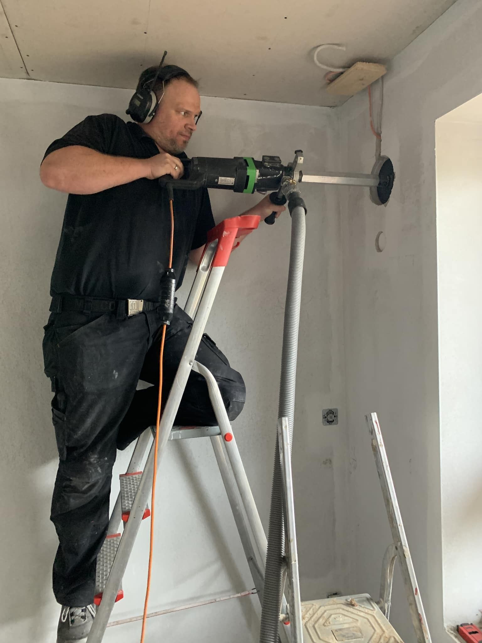 Ein Arbeiter bohrt von einer Leiter aus in einer unfertigen Wand in einem unfertigen Raum; Werkzeuge und Kabel liegen in der Nähe.