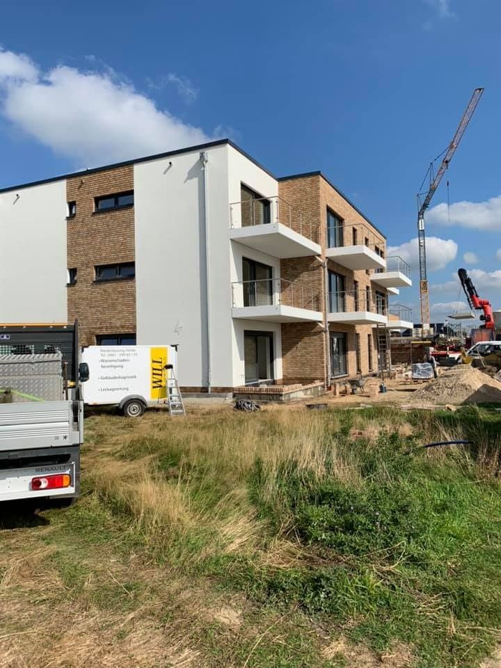 Ein teilweise errichtetes Mehrfamilienhaus mit Balkonen und Baumaschinen auf einer Rasenfläche unter blauem Himmel