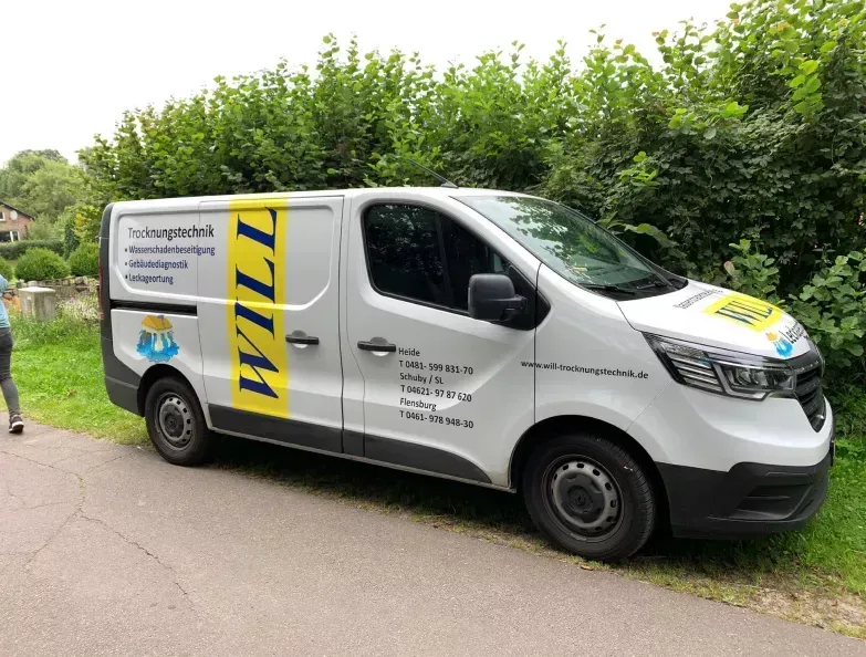 Ein weißer Lieferwagen mit gelbem „WILL“-Logo parkt am Straßenrand neben grünen Hecken.