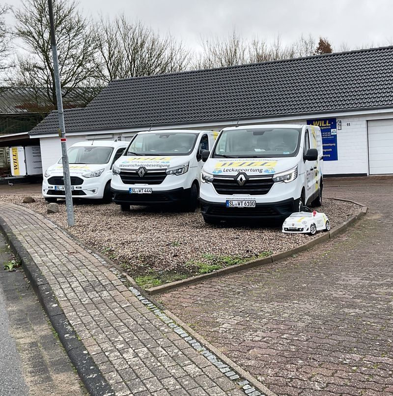 Zwei weiße Lieferwagen parkten vor einem Gebäude auf einem Schotterplatz, davor führte ein Kopfsteinpflasterweg.