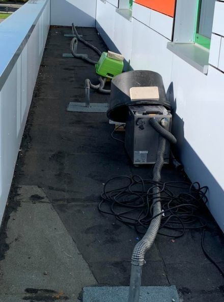 Ein Laufsteg auf dem Dach mit industriellen Lüftungsventilatoren, losen Kabeln und schwarzem Bodenbelag neben einer weißen Wand.
