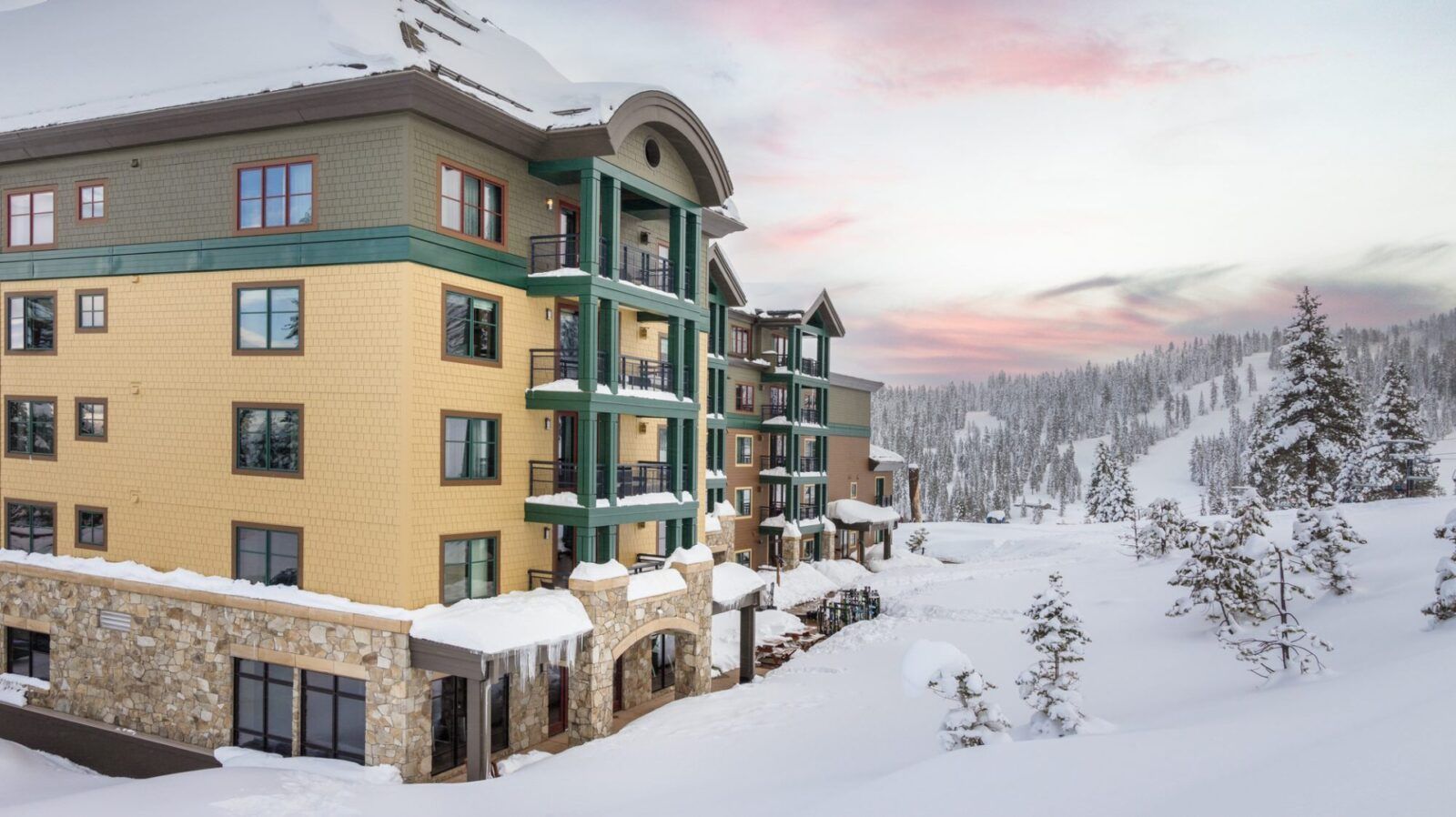 Snow-covered ski resort building with balconies; mountains and sunset in the background.