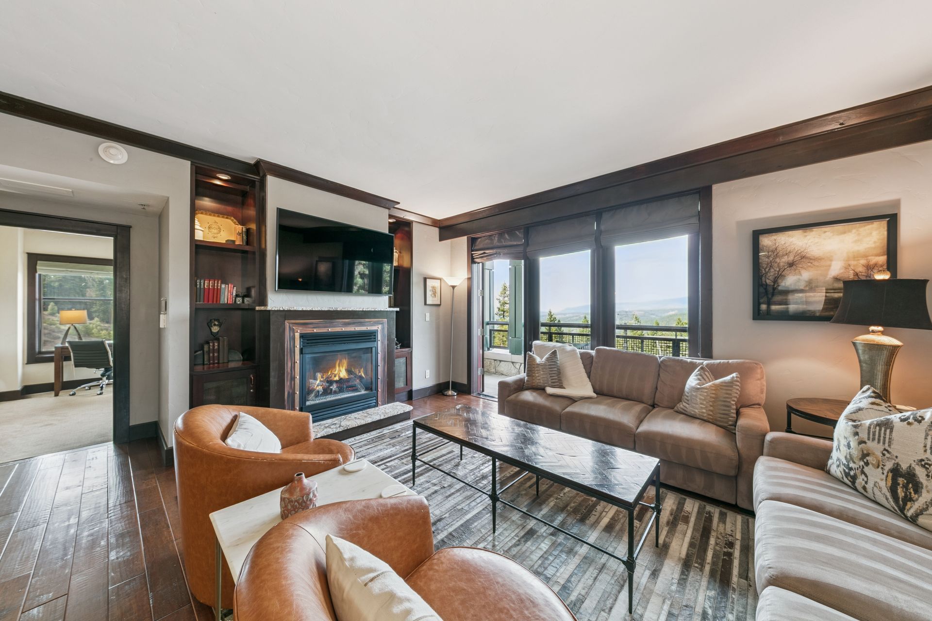 Living room with fireplace, large windows, and cozy seating. Dark wood trim, cityscape view.