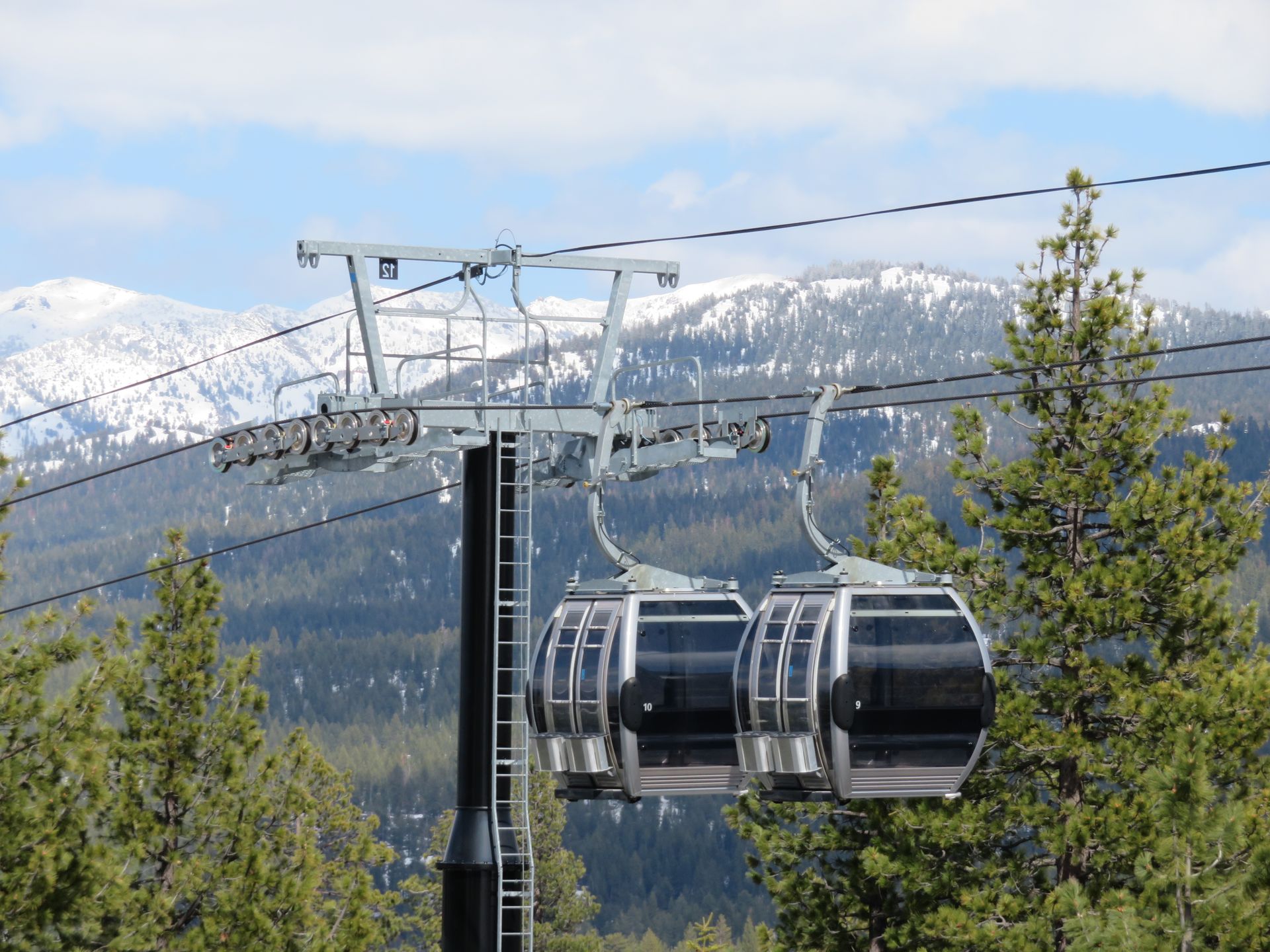 Gondola cars ascend against a backdrop of snow-capped mountains and trees under a partly cloudy sky.
