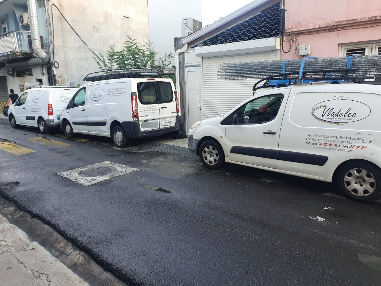 Des fourgonnettes de service blanches sont stationnées dans une rue étroite. Elles arborent toutes le même logo et certaines sont équipées d'échelles sur le toit.