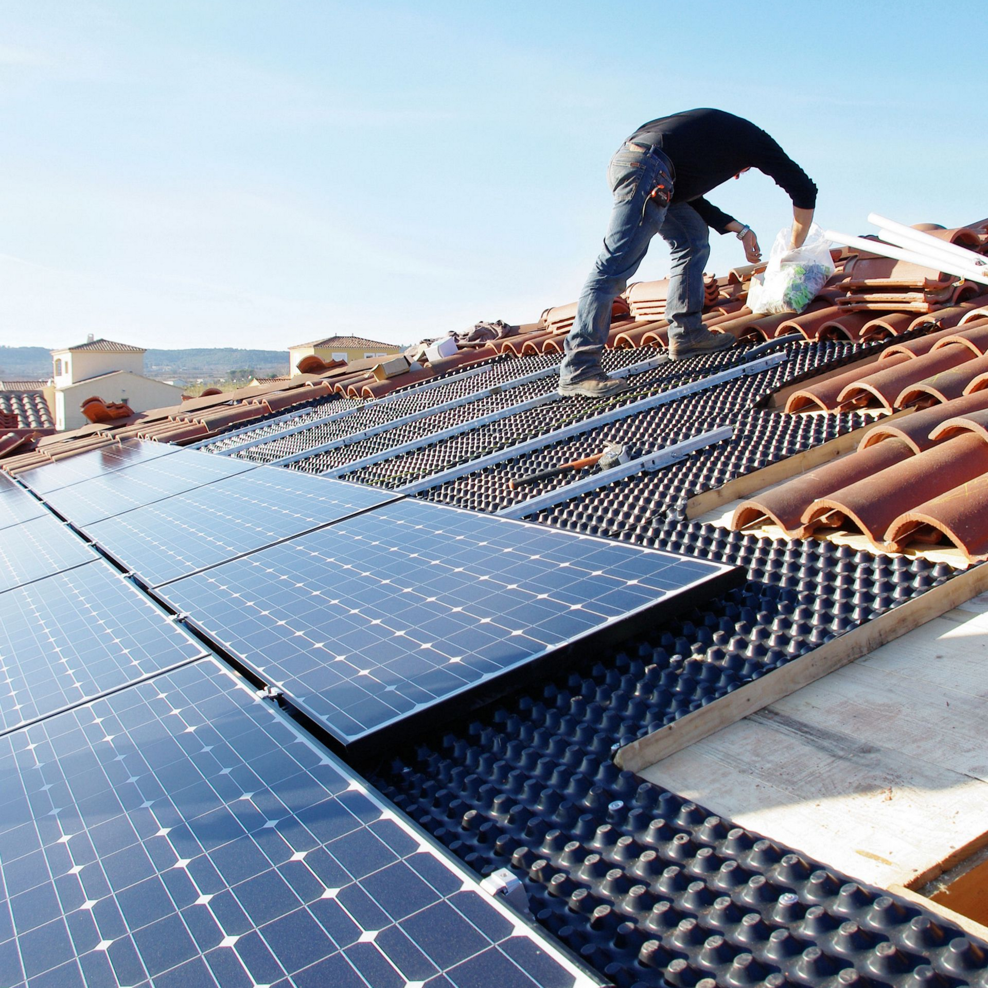 Un homme installe des panneaux solaires sur un toit de tuiles en terre cuite sous un ciel bleu azur.