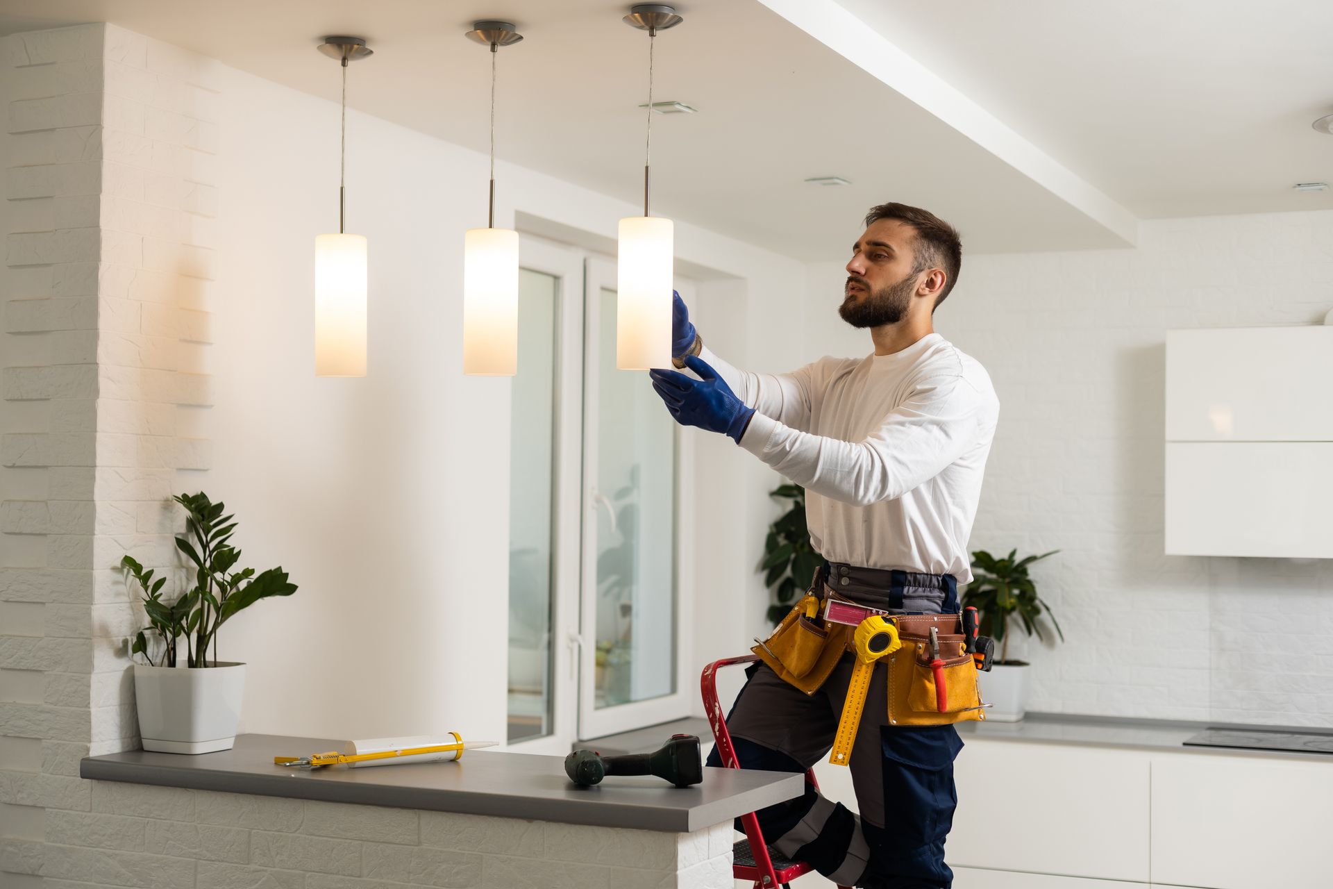 Un homme en tenue de travail, sur un escabeau, installe un luminaire dans une cuisine.